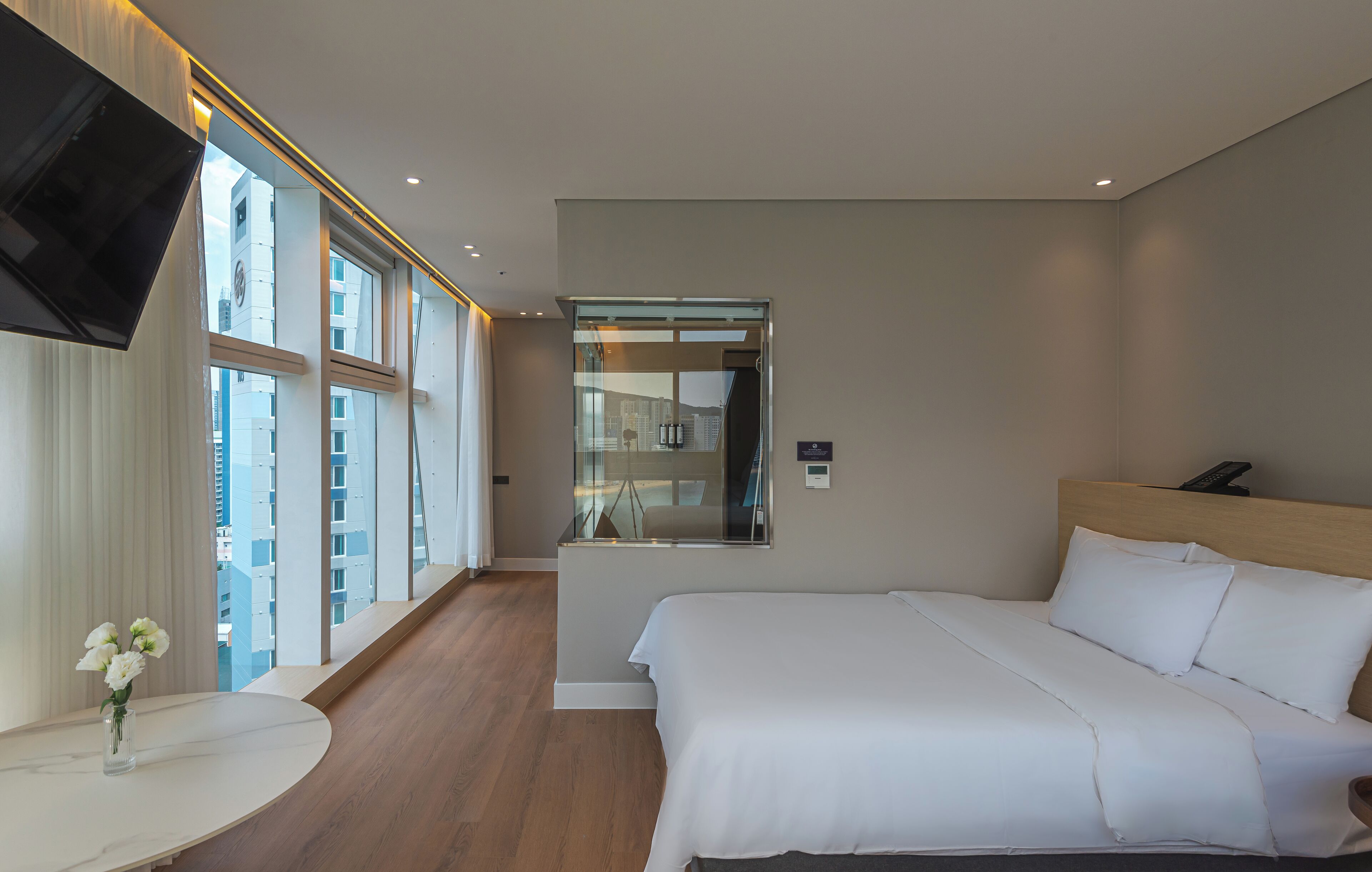 A modern hotel room with a large bed, a small round table with a vase of flowers, a TV mounted on the wall, and large windows offering a city view.
