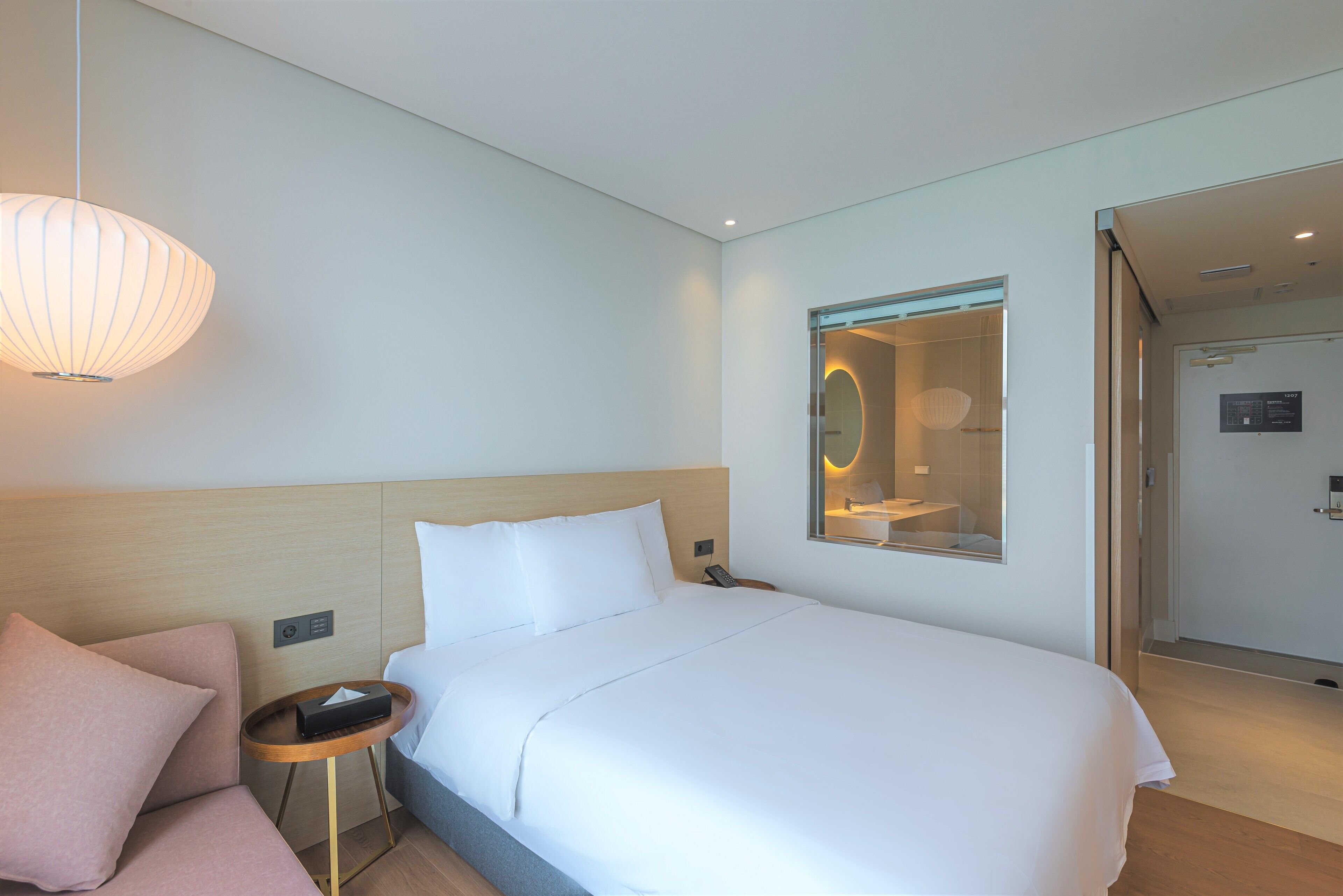 Habitación de hotel con cama, mesita de noche con teléfono y control remoto, una pequeña mesa redonda con lámpara y un espejo en la pared.