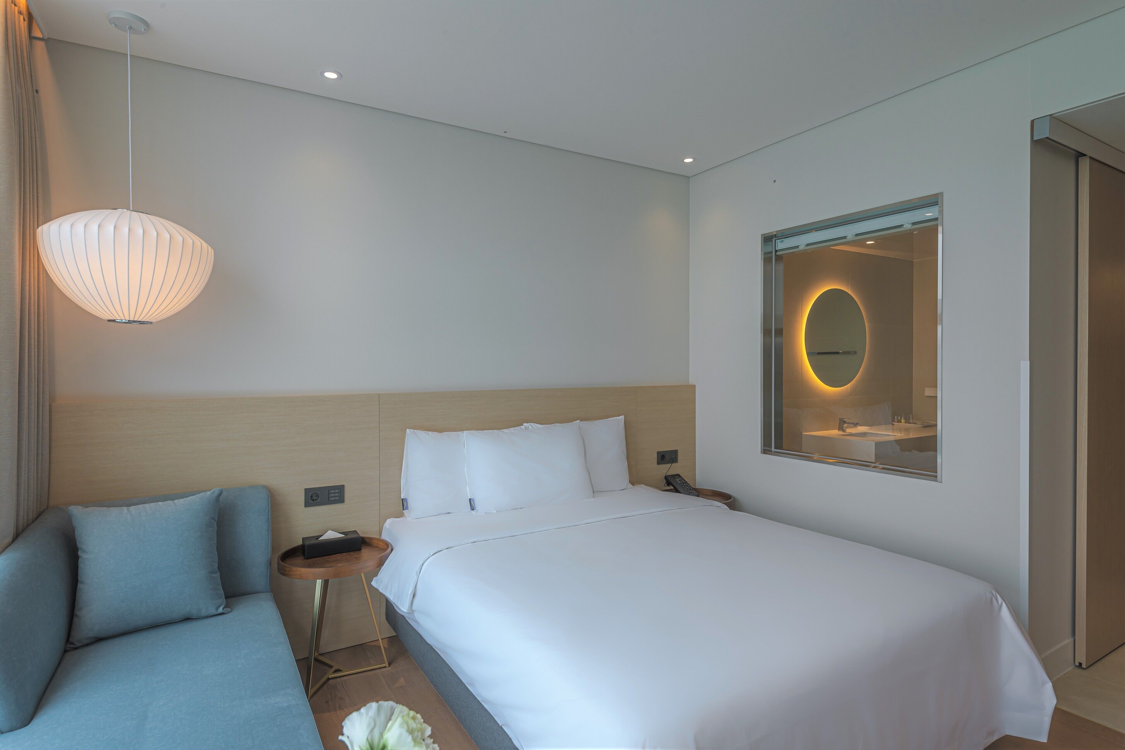 A modern hotel room with a bed, a sofa, a bedside table with a lamp, and a mirror with a circular frame.