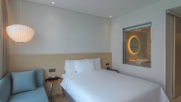 A modern hotel room with a bed, a sofa, a bedside table with a lamp, and a mirror with a circular frame.
