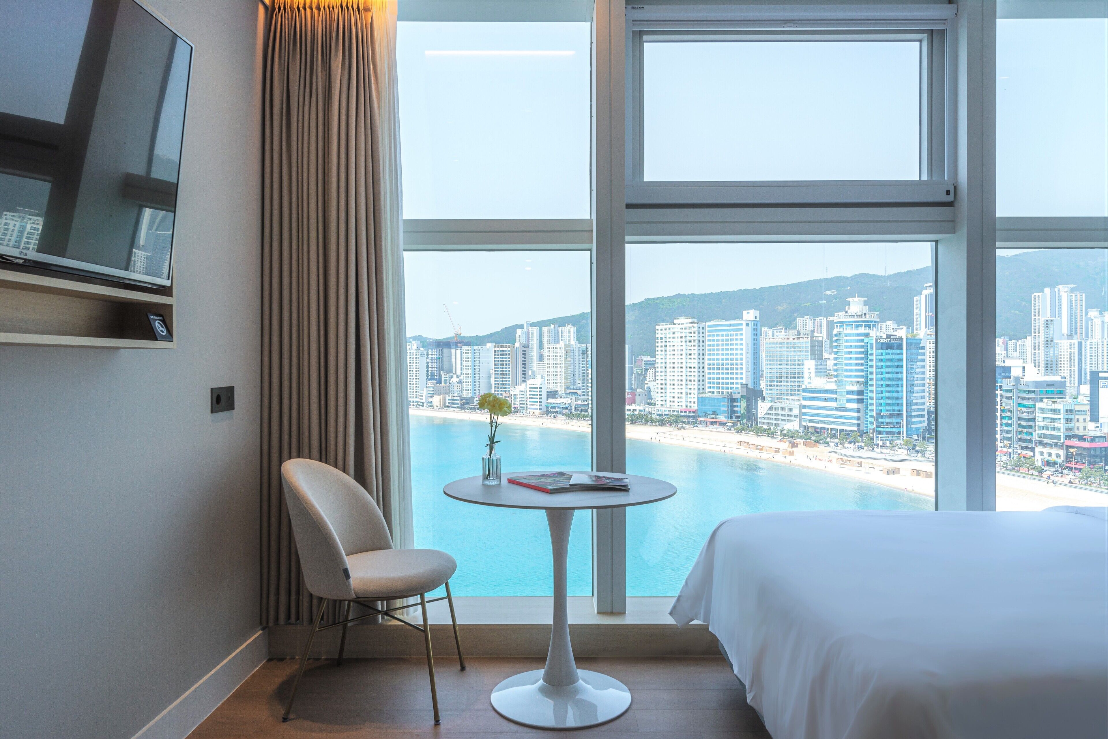 A hotel room with a bed, a chair, a small table, a TV, and a large window with a city view.