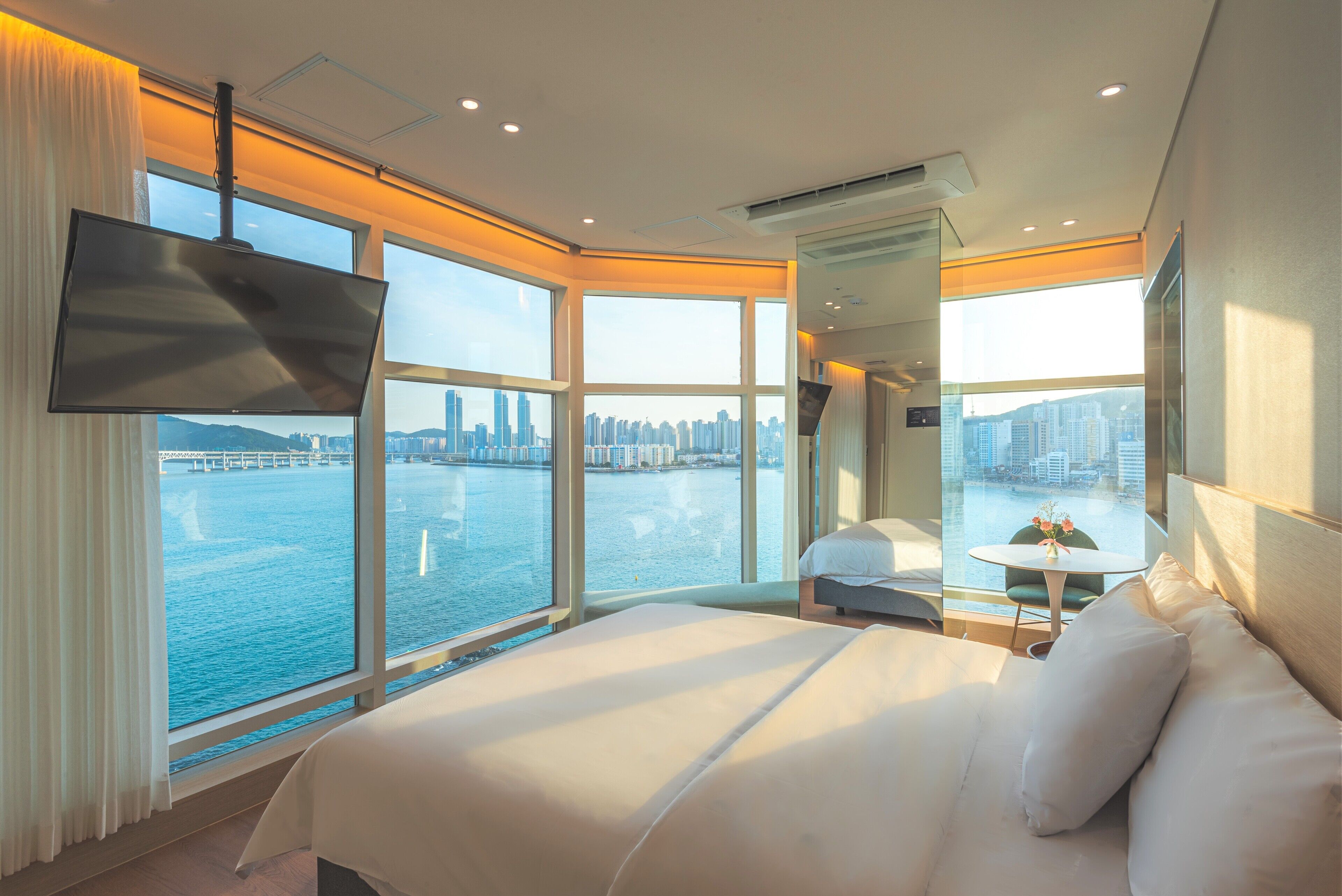 Een hotelkamer met een groot bed, een televisie, een klein tafeltje met een stoel en uitzicht op een stadsbeeld en water.