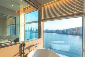 Un baño moderno con una bañera grande, un espejo y vistas a un horizonte urbano sobre una masa de agua.