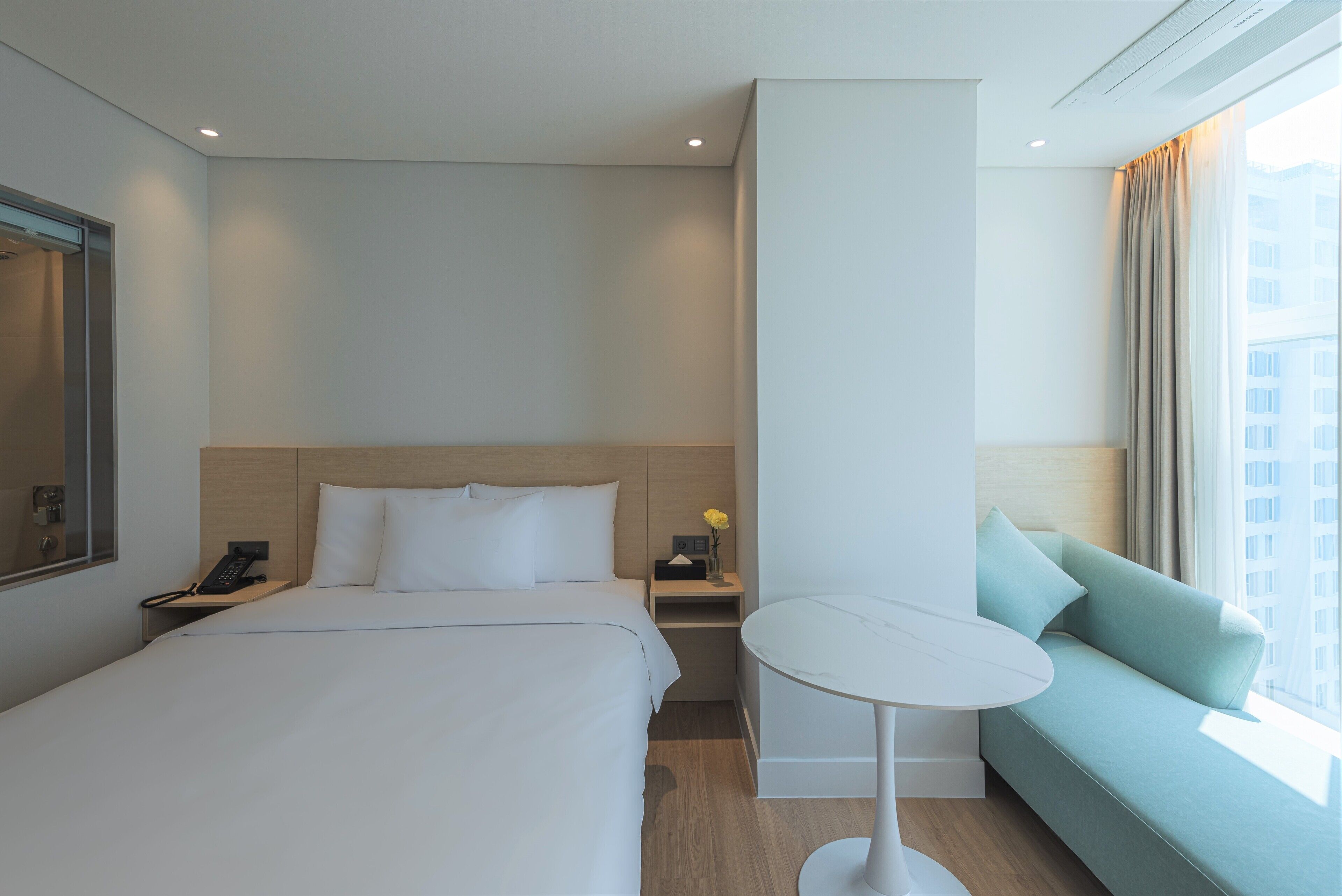 A modern hotel room with a large bed, a small bedside table, a bedside phone, a round table, and a sofa.