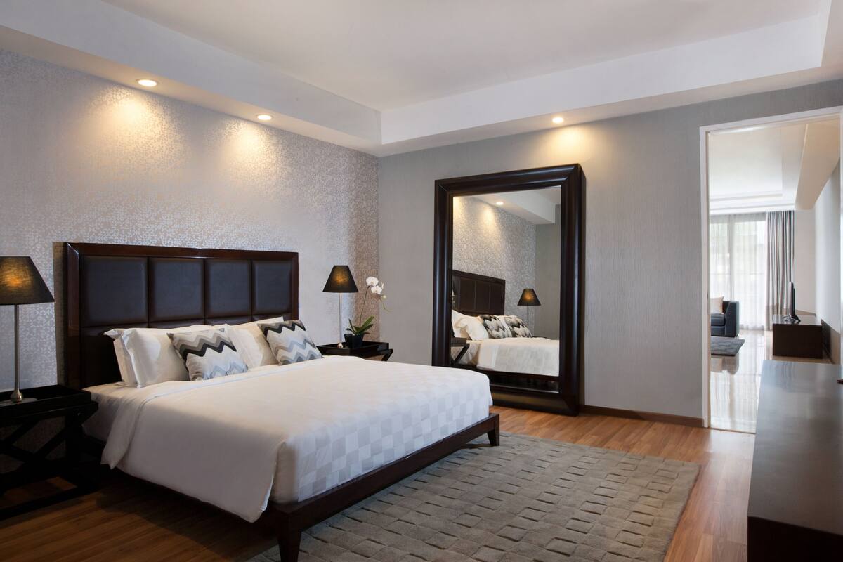 A modern hotel room with a large bed, two bedside lamps, a mirror, and a view into the adjacent living area.
