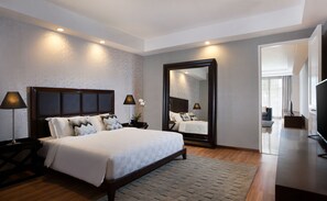 A modern hotel room with a large bed, two bedside lamps, a mirror, and a view into the adjacent living area.