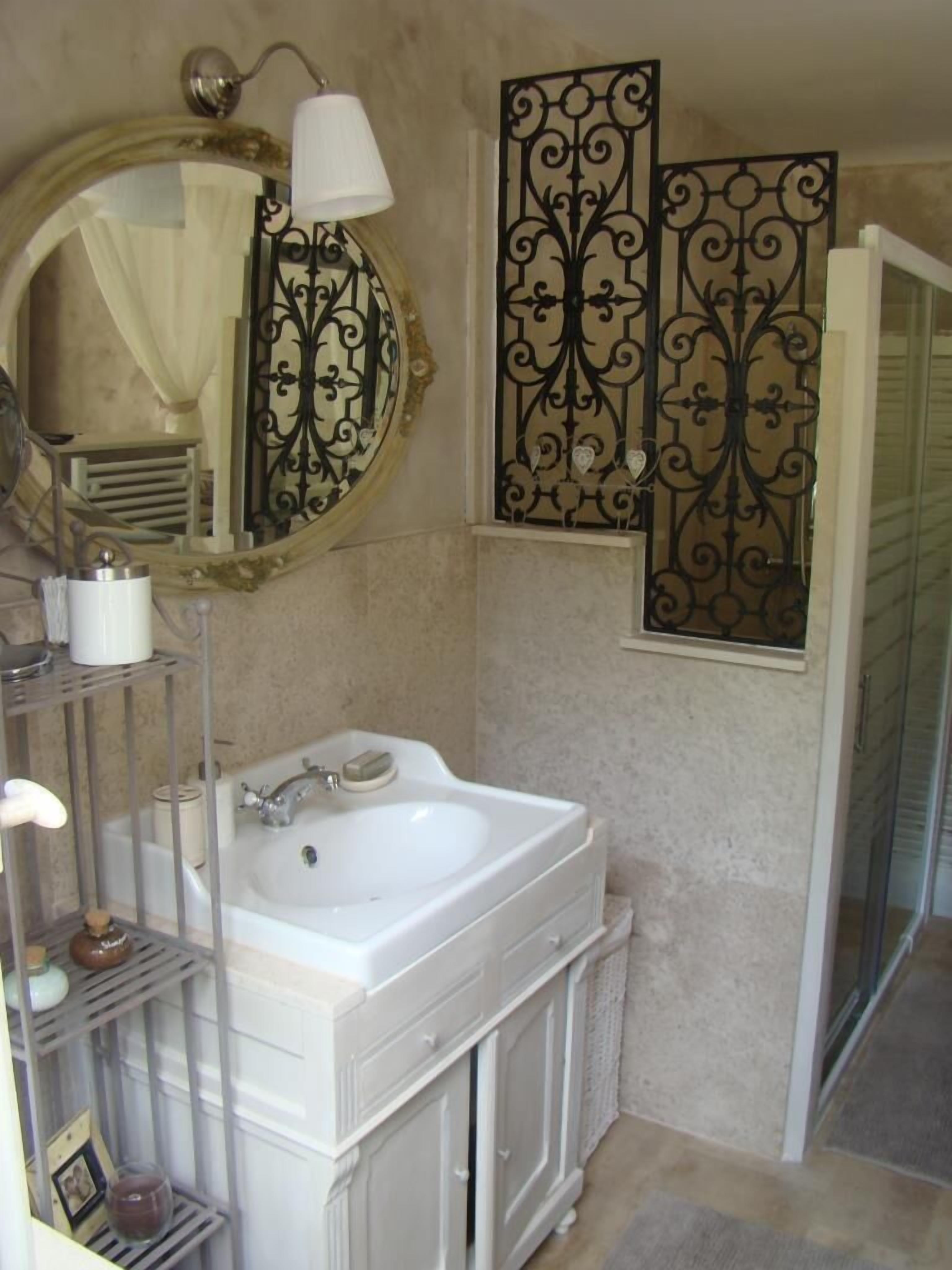 Une salle de bain avec un lavabo blanc, un meuble-lavabo et un miroir. Un panneau décoratif en métal se trouve au-dessus du miroir, et une porte de douche est située à droite.
