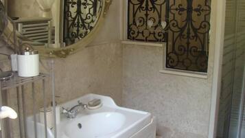 Une salle de bain avec un lavabo blanc, un meuble-lavabo et un miroir. Un panneau décoratif en métal se trouve au-dessus du miroir, et une porte de douche est située à droite.