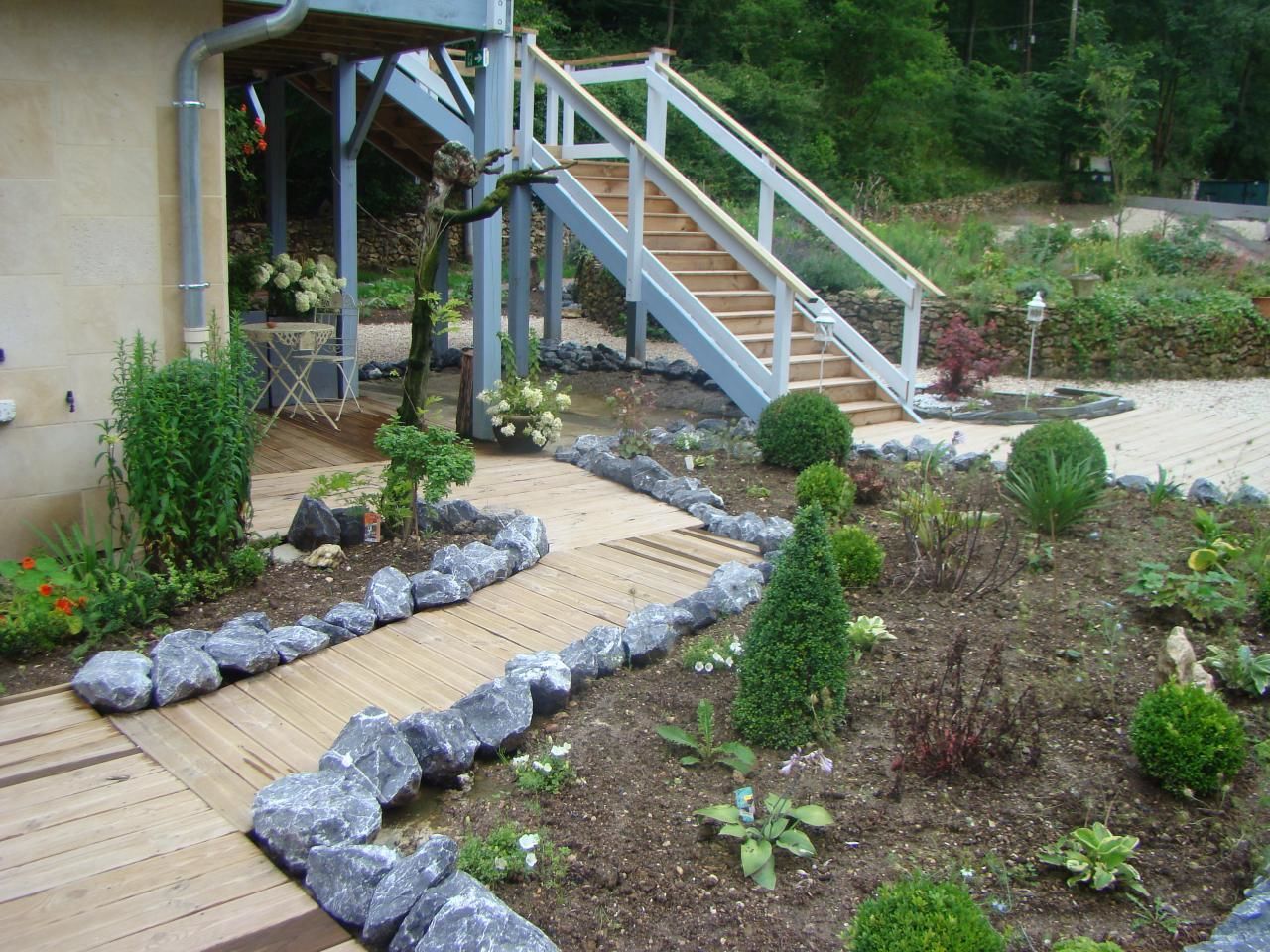 Ein Garten mit Holzterrasse, Steinmauer und einer Treppe zu einem überdachten Bereich.