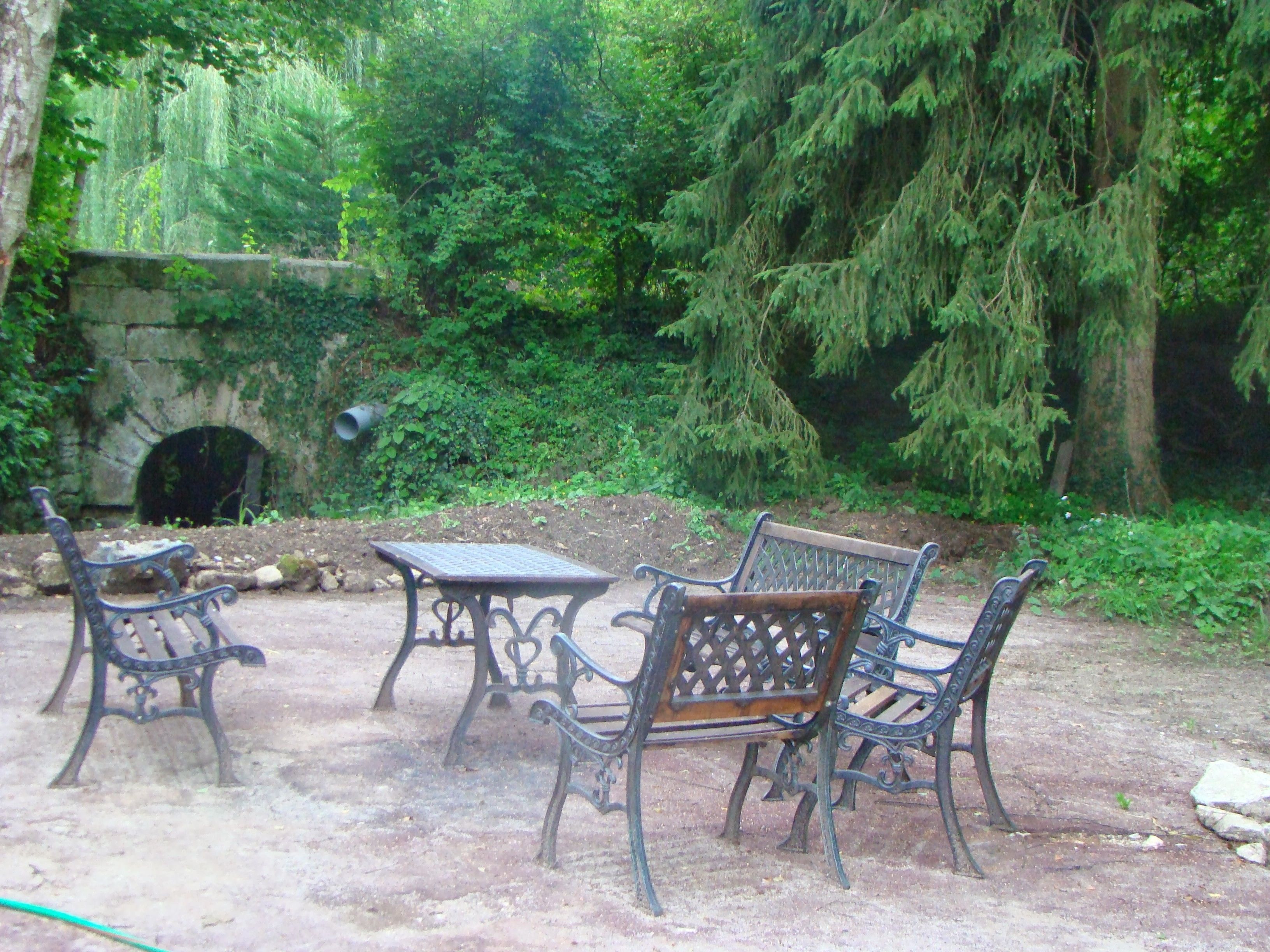 An outdoor seating area with wrought iron tables and benches, surrounded by lush greenery and a stone wall.