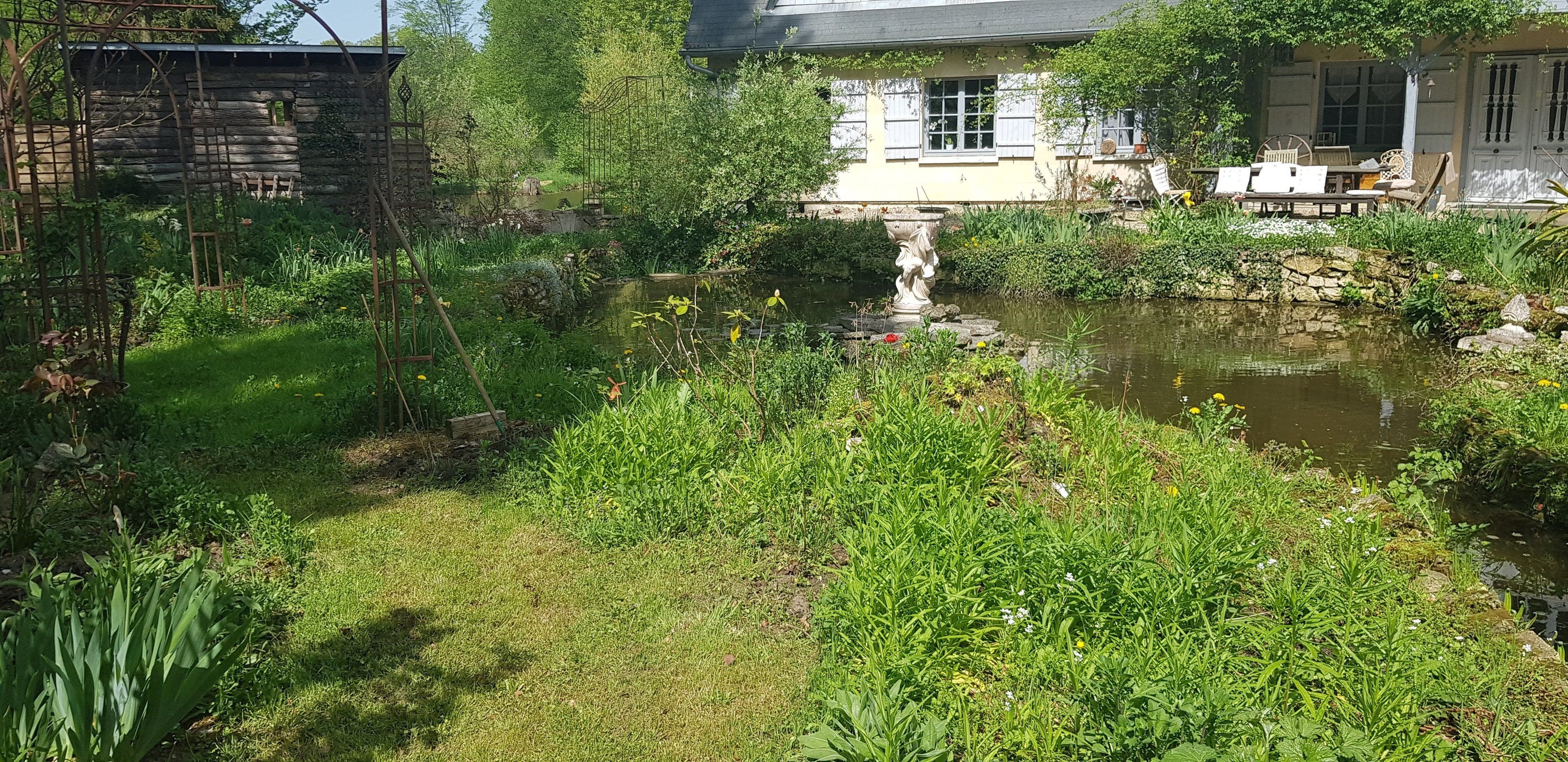 Een tuin met een vijver, een rustieke stenen muur en een gebouw met een witte gevel en luiken.