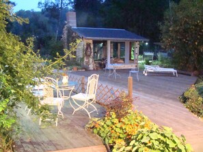 Ein überdachter Außenbereich mit Tischen und Stühlen, umgeben von Pflanzen und einer Holzterrasse.