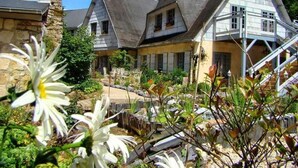 Une maison à deux étages avec une cheminée en pierre, une terrasse en bois et un jardin agrémenté de fleurs blanches.