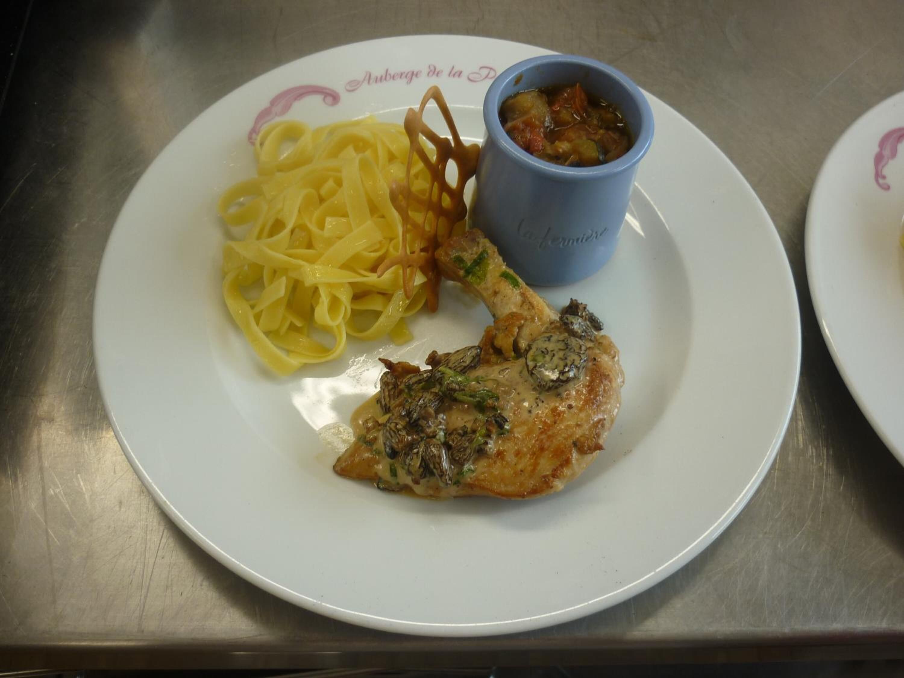 Een bord met een stukje kip, fettuccine en een klein blauw kopje saus.