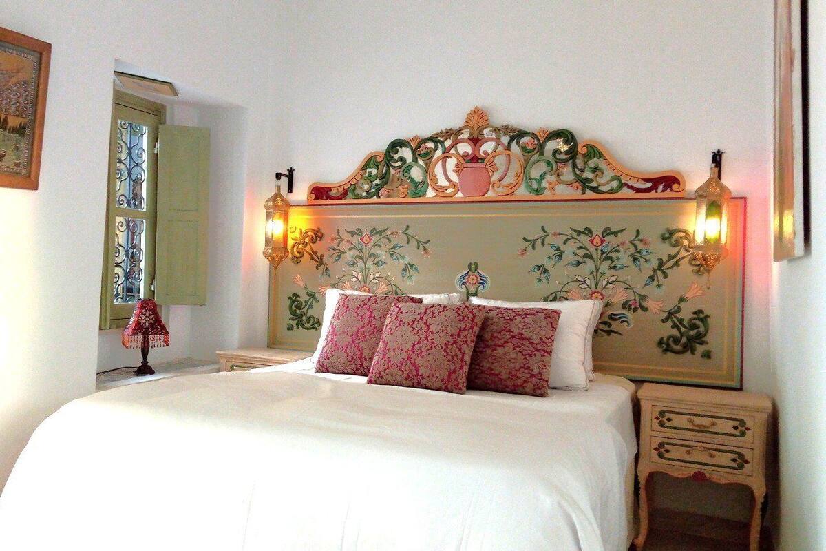 A bedroom with a large bed, ornate headboard, two wall-mounted lamps, and a nightstand with drawers.