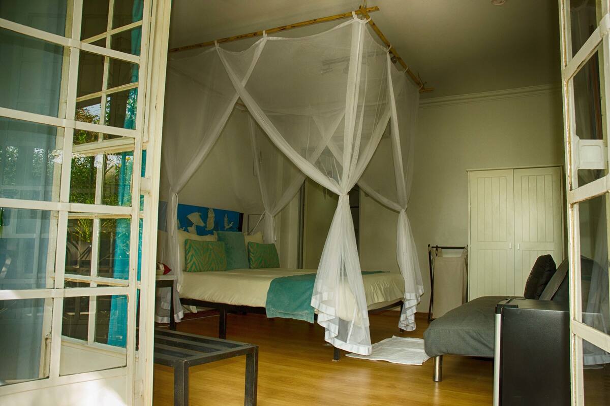 A room with a bed under a white canopy, a bedside table, a chair, and a view of the outside through a window.