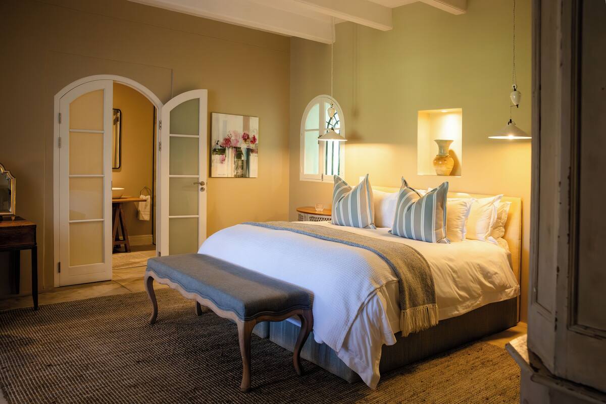 A bedroom with a large bed, a bench, a bedside table, a mirror, and a window with a view.