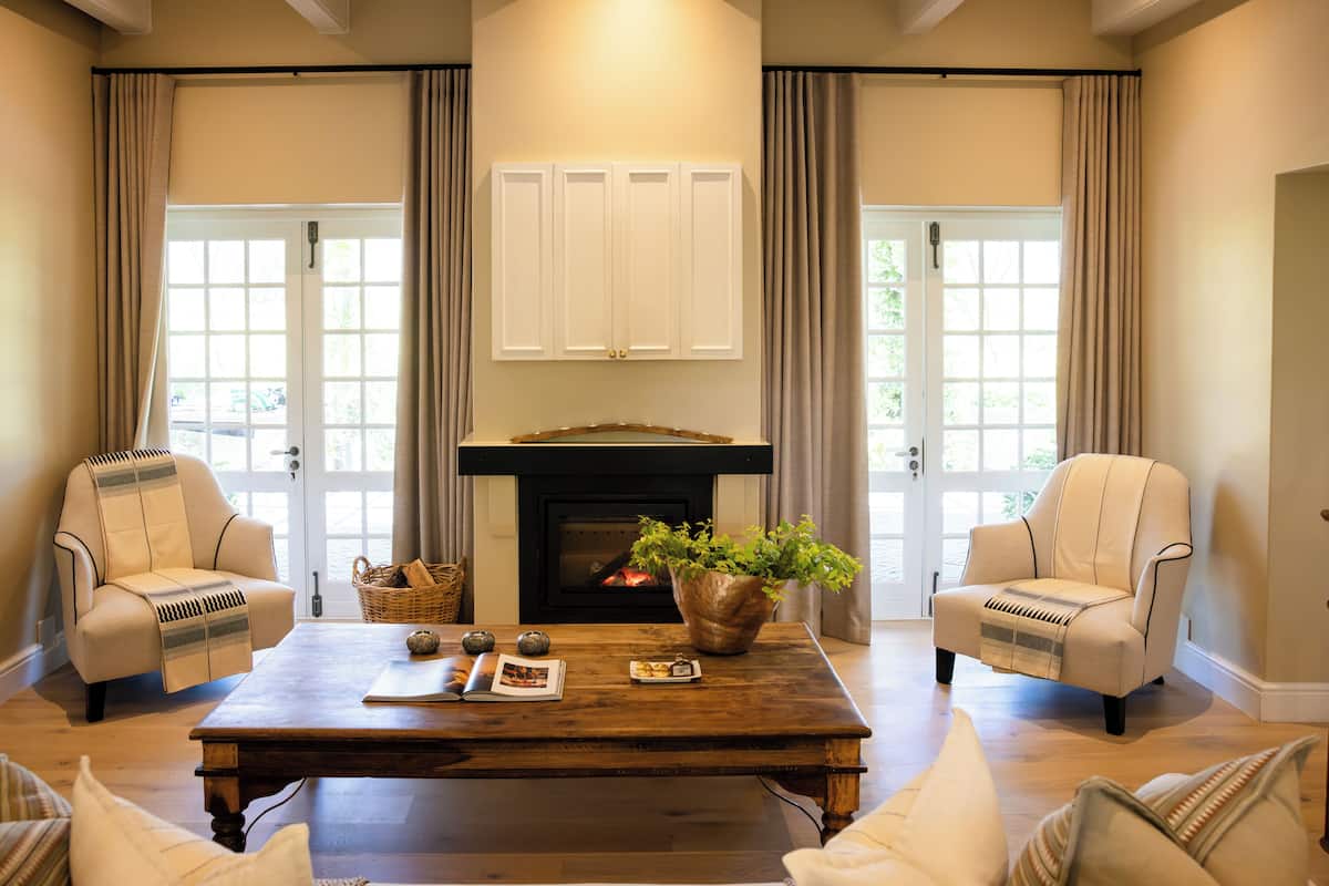 A living room with a fireplace, two armchairs, a coffee table, and a large window with curtains.