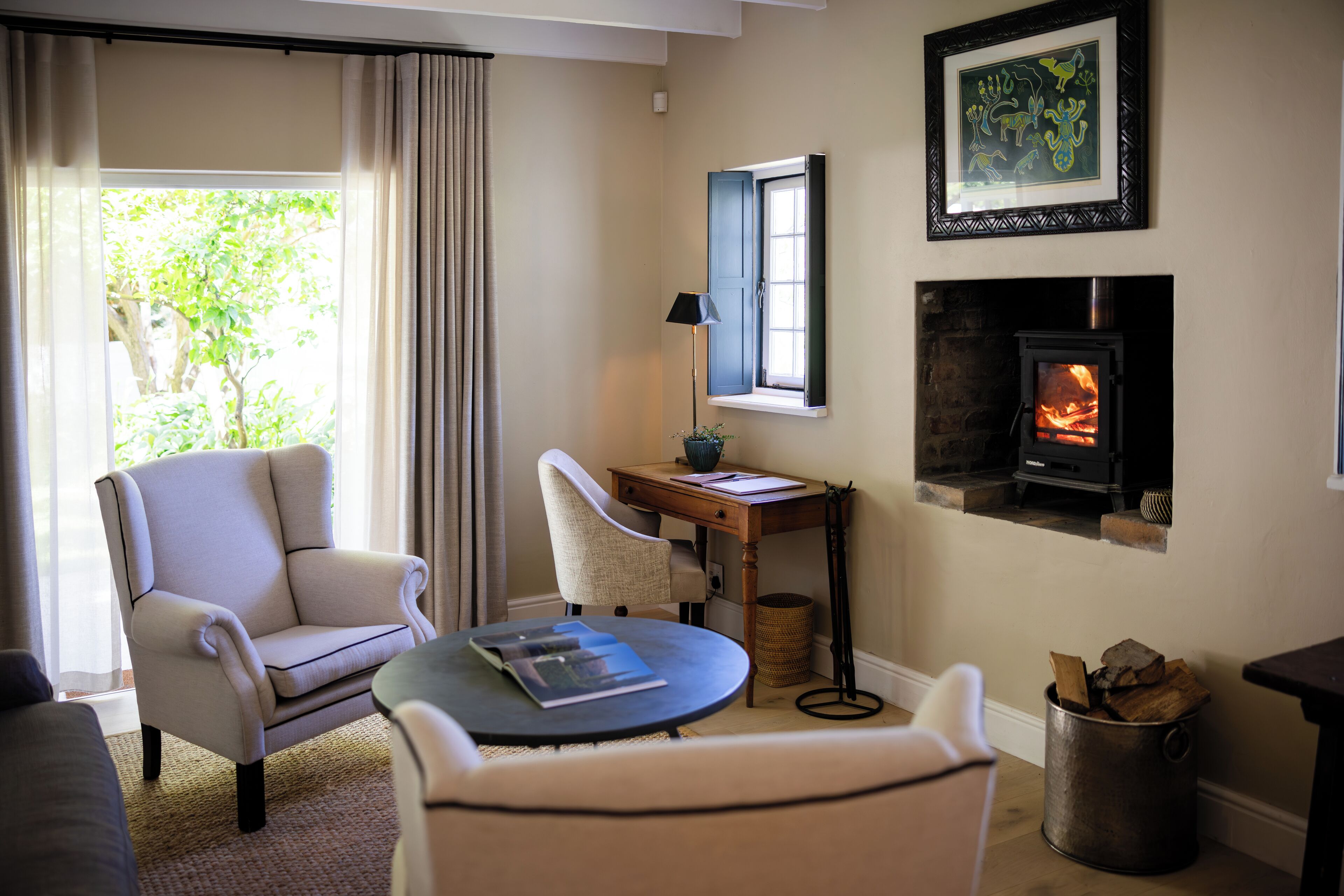 Una sala de estar acogedora con chimenea, un sillón cómodo, una mesita con lámpara y vistas al exterior con vegetación.