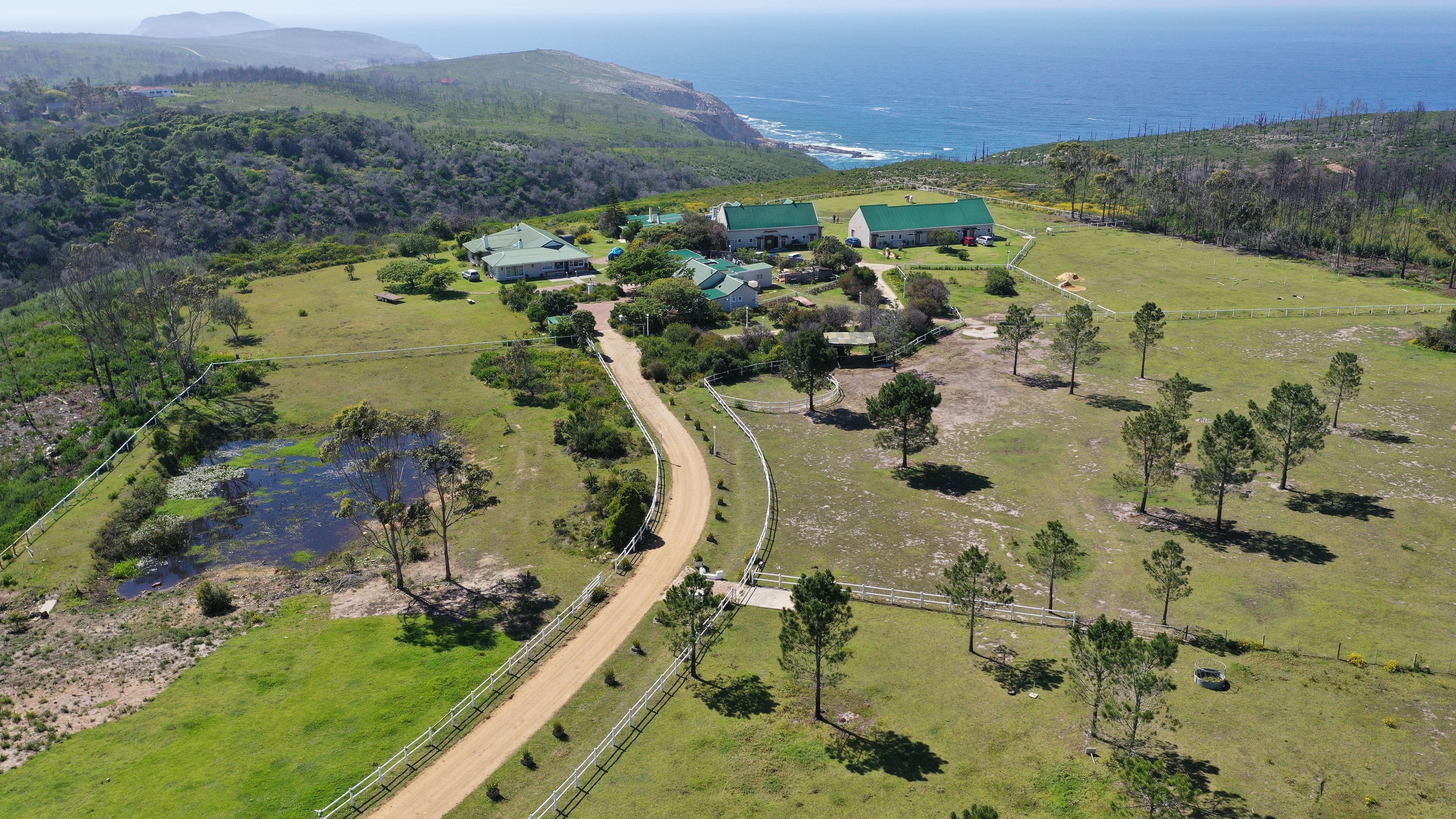 An aerial view of a coastal property with a winding road, greenhouses, and a fenced pasture.