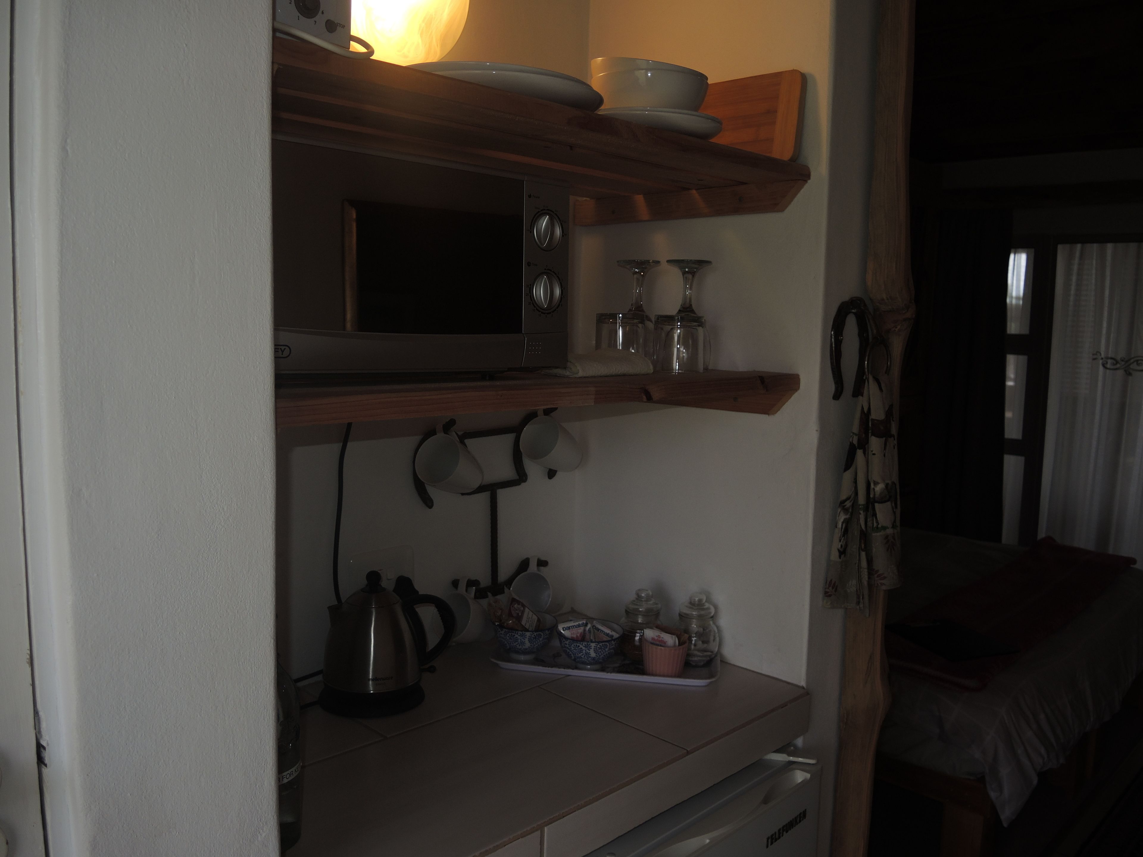A compact kitchen with a microwave, kettle, and shelves holding dishes and jars.
