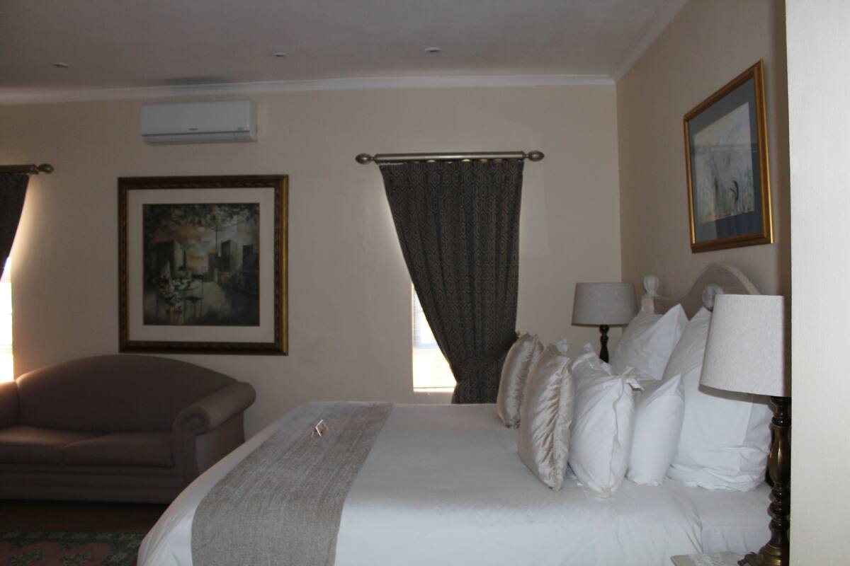 A hotel room with a bed, a chair, a painting, a window with curtains, and a lamp.