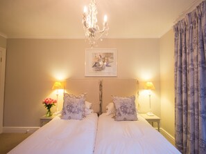 A double bed with a chandelier above, two bedside lamps, a framed picture on the wall, and a window with curtains.