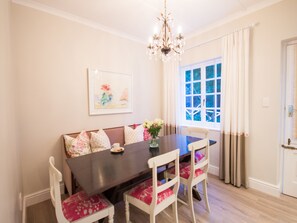 A dining area with a dark wooden table, white chairs with red cushions, a chandelier, a framed picture on the wall, and a window with curtains.