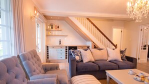 Een moderne woonkamer met een bank, een gestreept voetenbankje en een salontafel. Er is een trap die leidt naar een hoger gelegen niveau, met een zichtbare keuken.