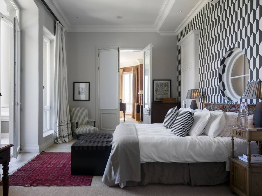 Une chambre avec un grand lit, une table de chevet en bois, un bureau et une chaise. Il y a une fenêtre avec des rideaux, un tapis rouge et un mur à motifs.