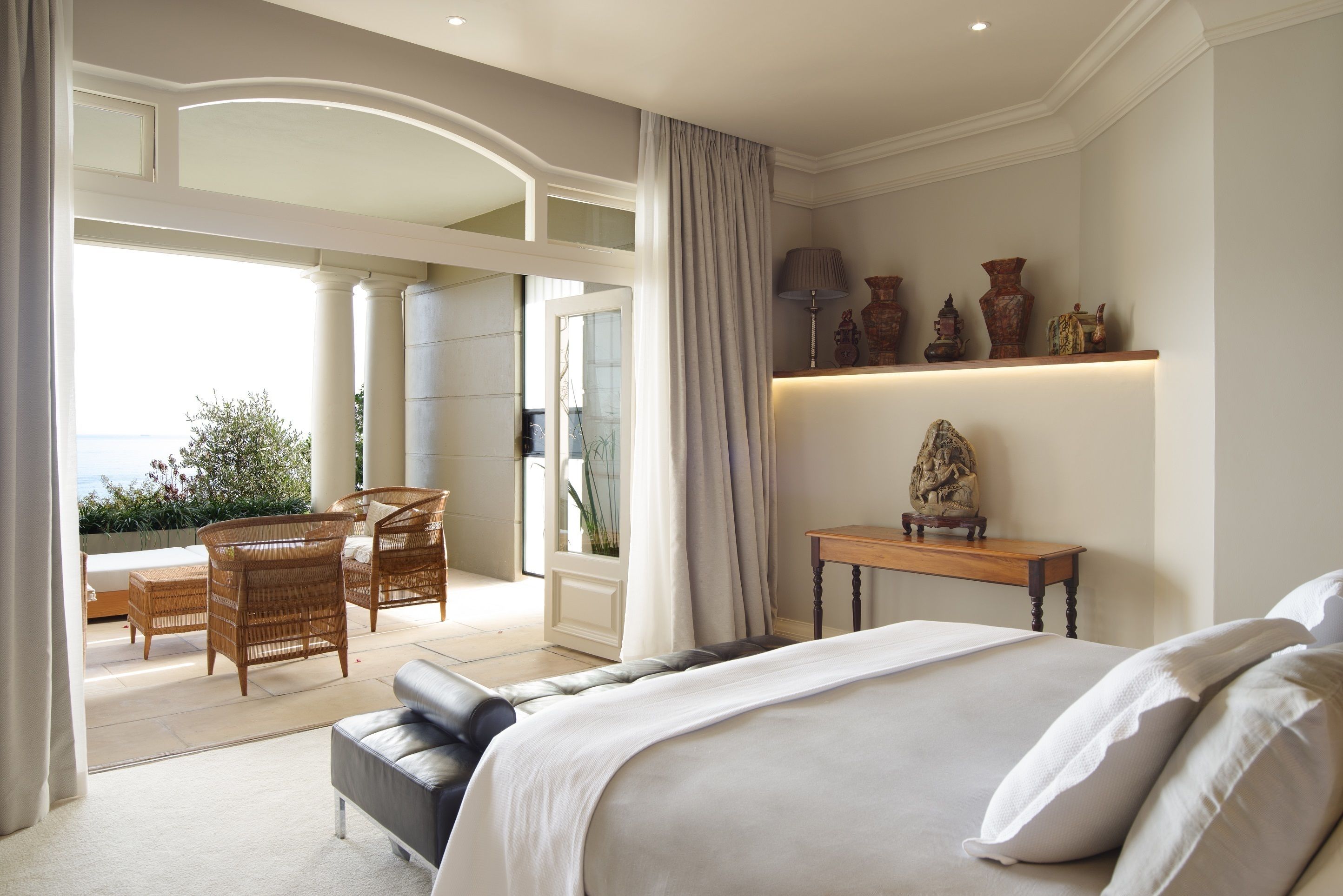 A bedroom with a large bed, a small table with decorative items, a sofa, and a balcony with wicker furniture.