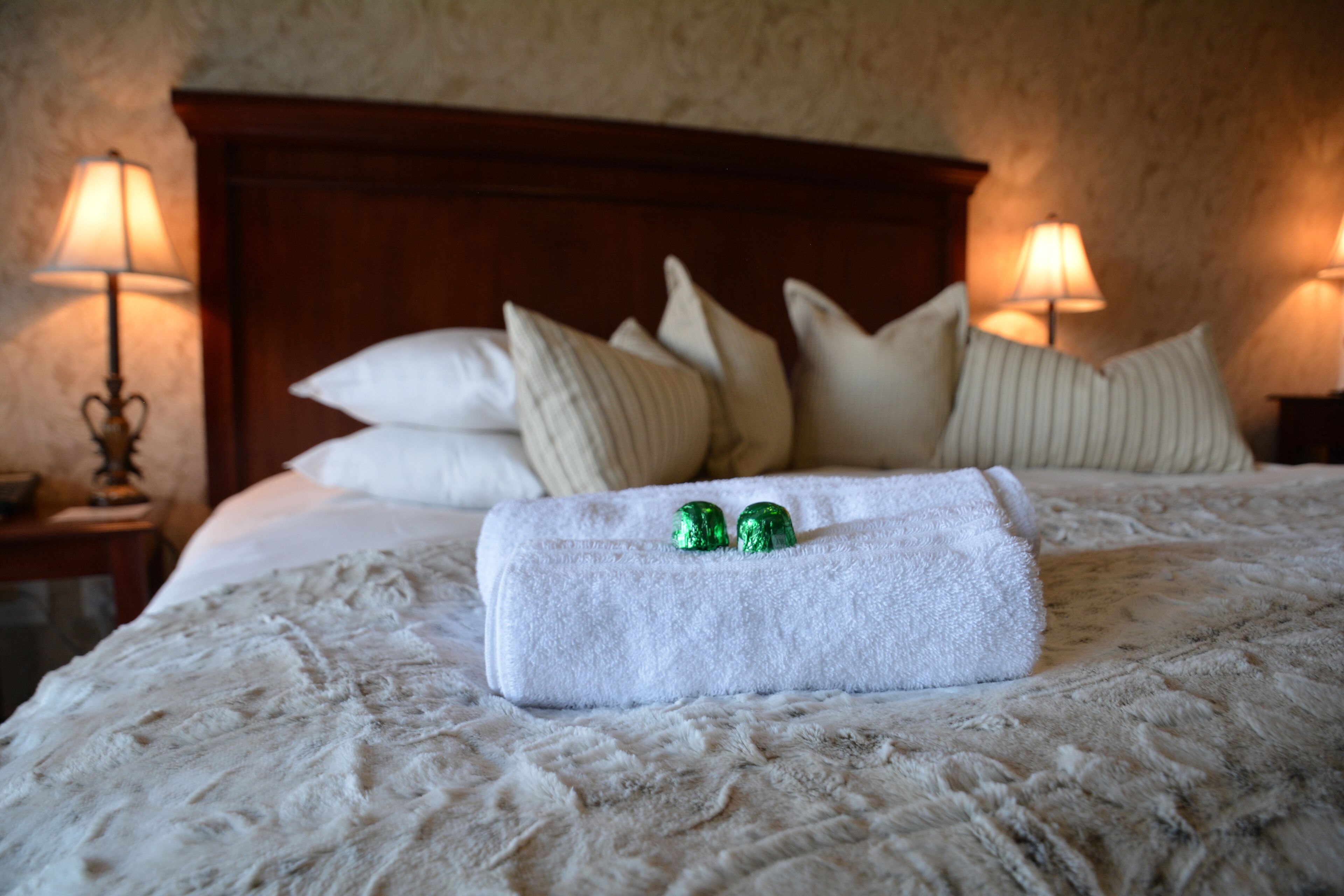 A neatly made bed with a white towel and two green bottles, flanked by two bedside lamps and a wooden headboard.