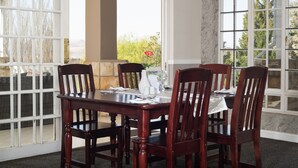 Un coin repas avec une table et des chaises en bois, de grandes fenêtres donnant sur un paysage verdoyant et un vase contenant une seule fleur rouge.