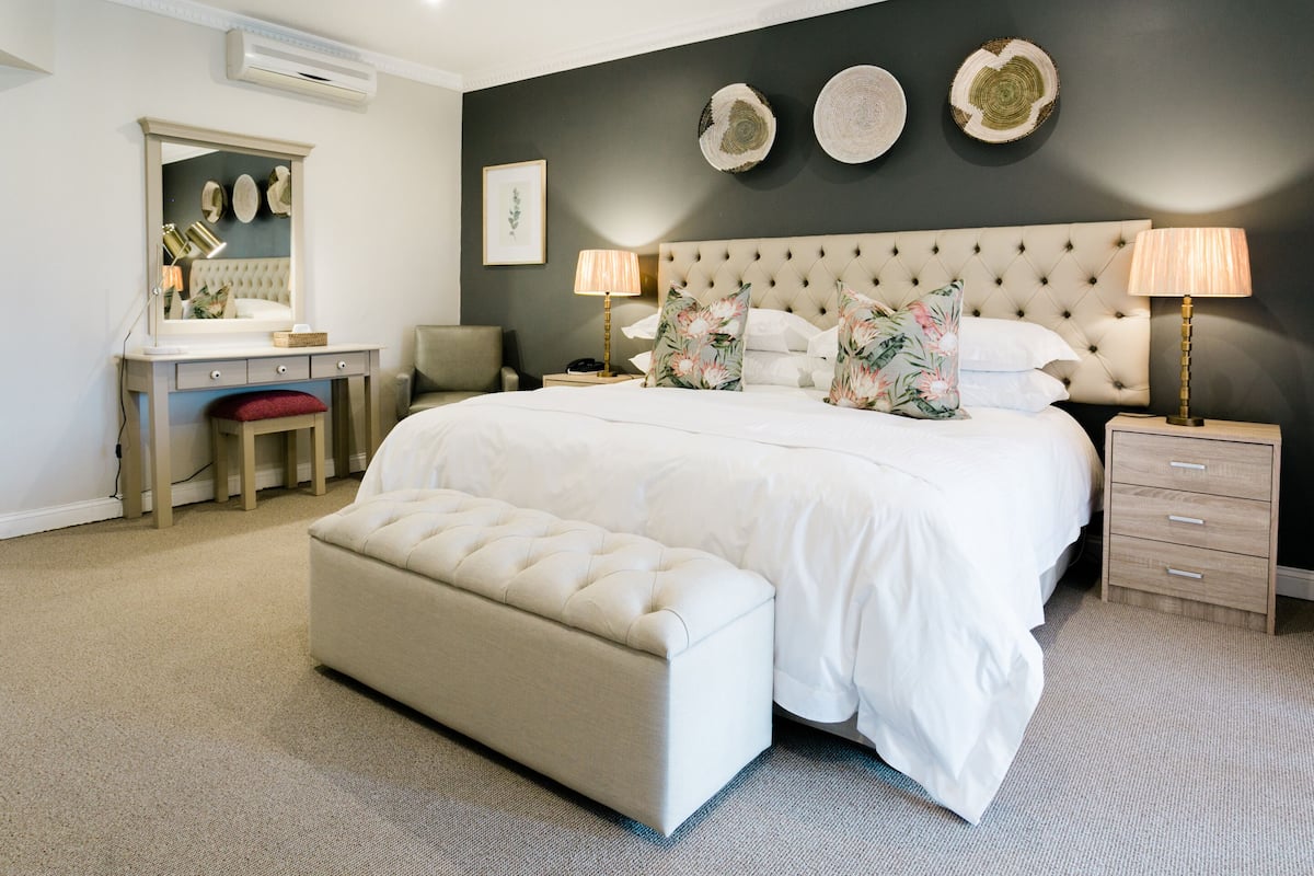 A bedroom with a large bed, a bedside table, a nightstand, a dresser, a mirror, and a bench.