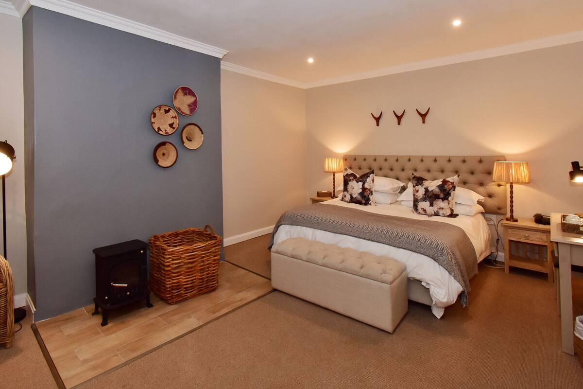 A bedroom with a large bed, a nightstand, a lamp, a wicker basket, and a fireplace.