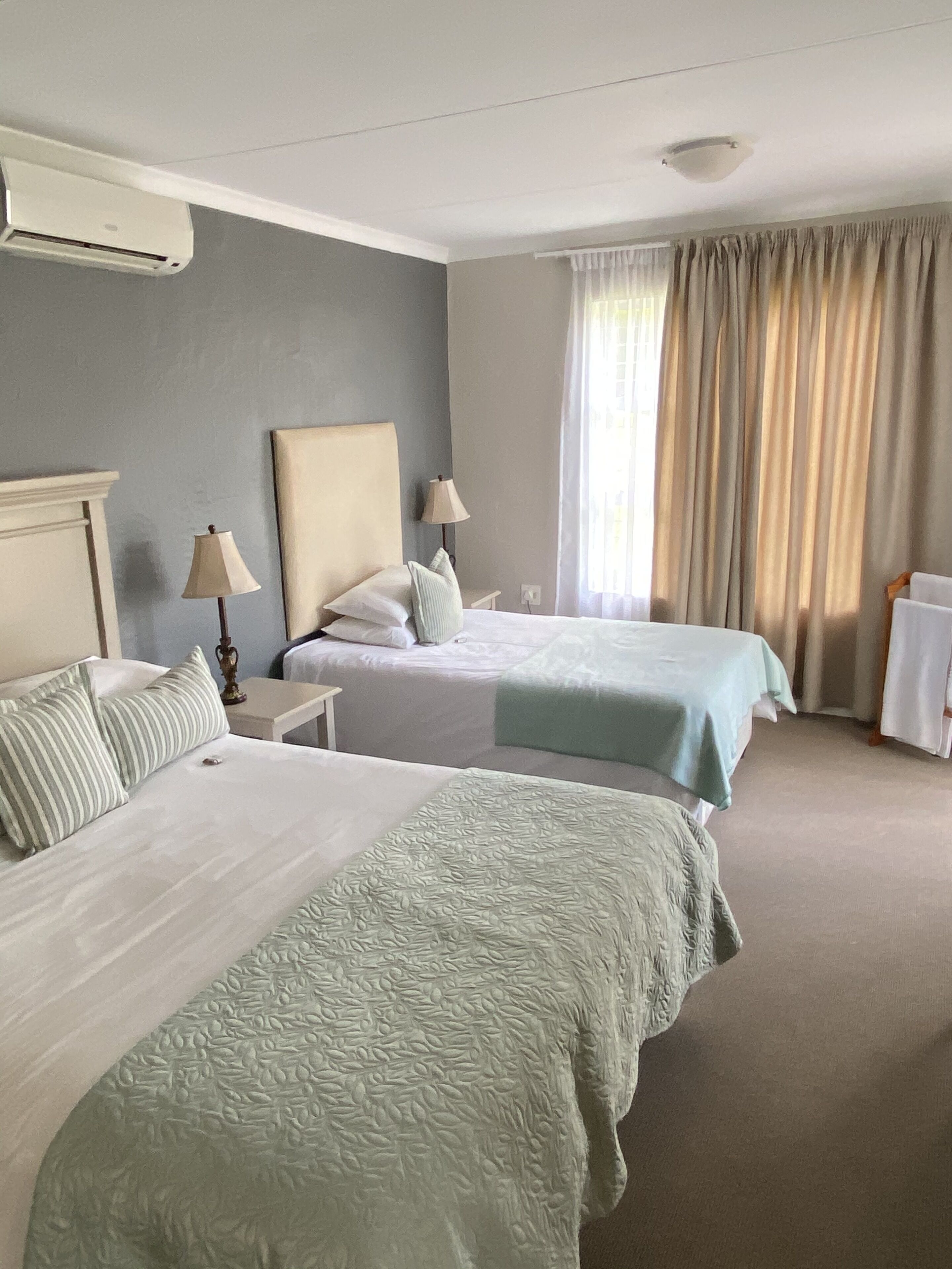 A hotel room with two beds, a ceiling fan, a window with curtains, and a wall-mounted air conditioning unit.