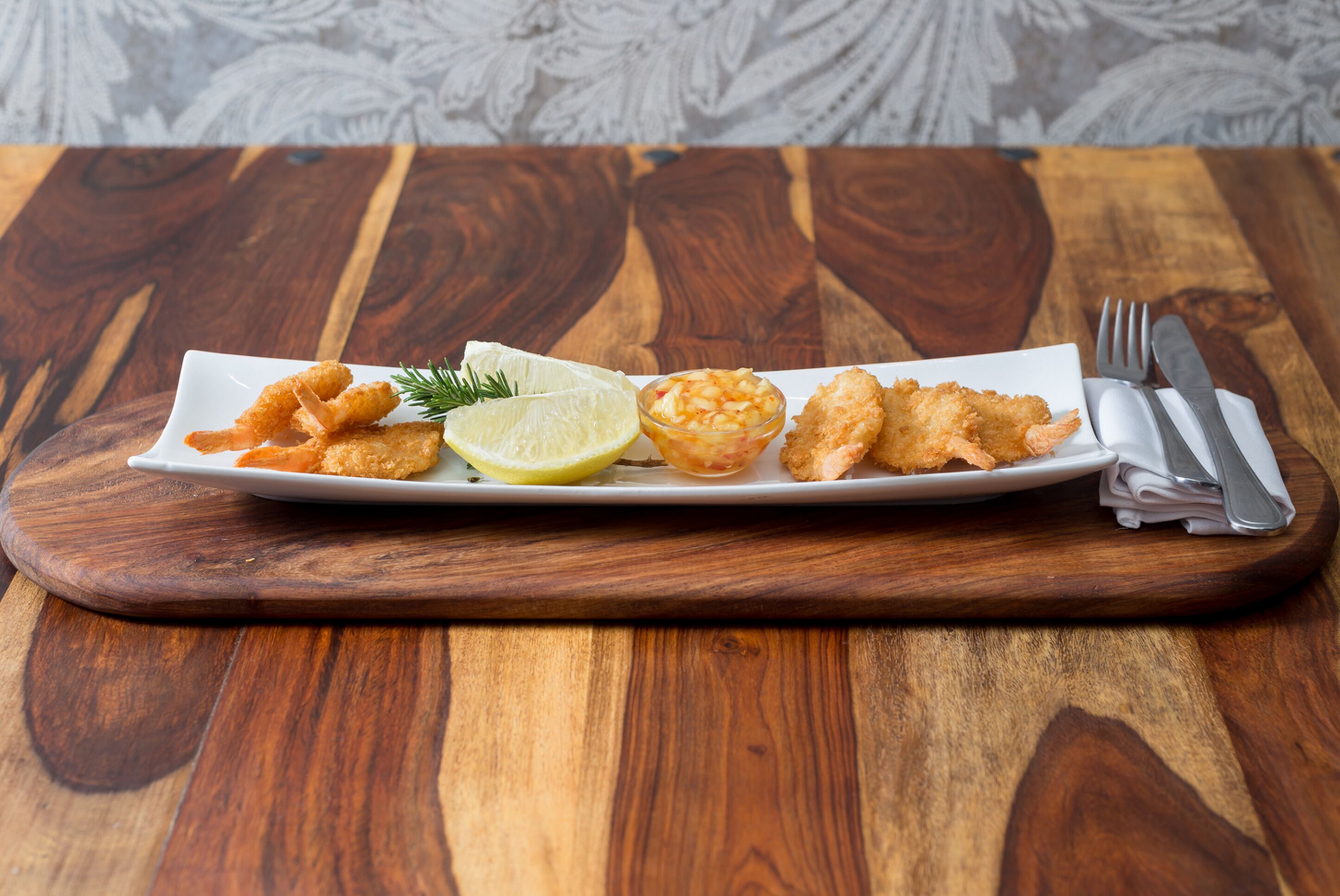 A white rectangular plate with fried food items, a lemon wedge, and a small bowl of sauce on a wooden table.