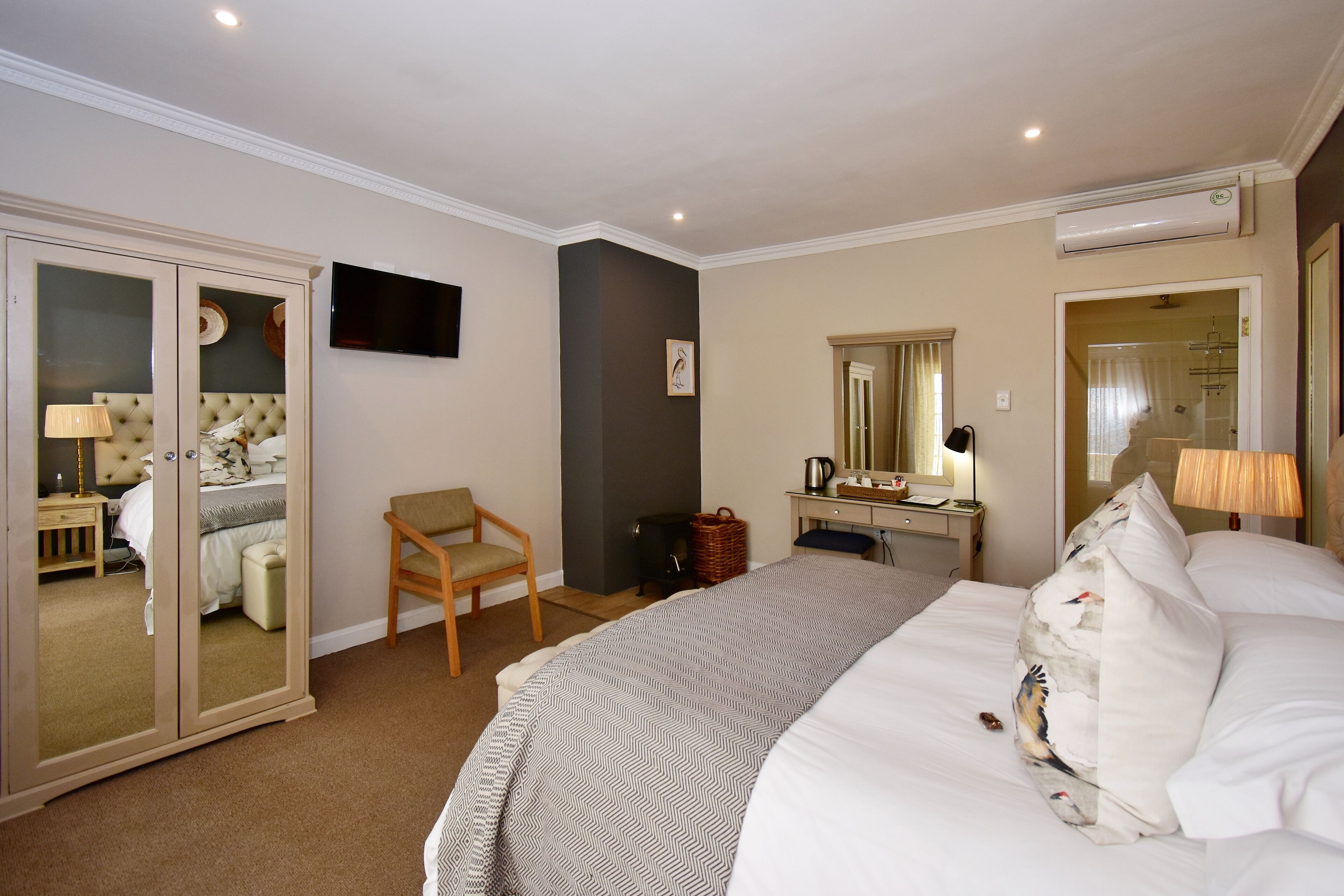 A hotel room with a bed, a chair, a desk with a lamp, a mirror, and a wardrobe with a TV.