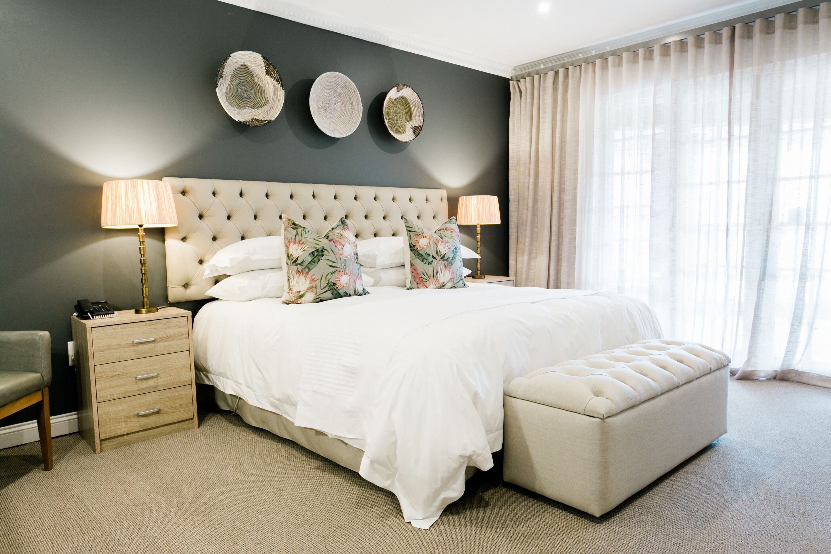 Une chambre à coucher avec une tête de lit capitonnée, un grand lit agrémenté d’oreillers à motifs floraux, une table de chevet avec des lampes et une grande fenêtre avec des rideaux.
