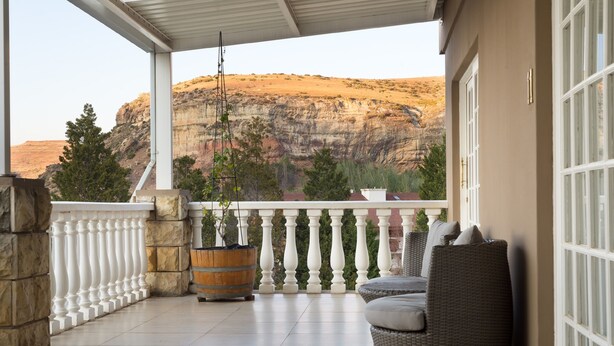 Un balcon abrité offrant une vue sur un paysage montagneux, avec des piliers en pierre, un canapé en osier et un jardinière en bois.