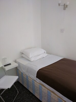 A small, neatly made bed with white bedding and a brown blanket, a white bedside table with a remote control, and a wall-mounted light fixture. - ABC Bed & Breakfast (London)