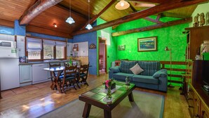 A cozy living room with a green wall, wooden ceiling beams, a dining area with a table and chairs, and a kitchen with white cabinets and a microwave. - Apartamentos Rurales Balcón Real (Teverga)