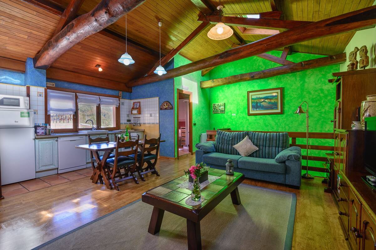 A cozy living room with a green wall, wooden ceiling beams, a dining area with a table and chairs, and a kitchen with white cabinets and a microwave.