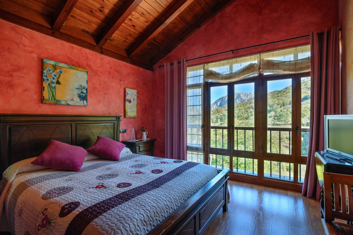 A bedroom with a large bed, wooden headboard, and a view of a mountainous landscape through a large window.