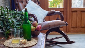 A wooden rocking chair with a cushion, a woven placemat with a glass of liquid, a green bottle, and a bowl of nuts. - Apartamentos Rurales Balcón Real (Teverga)