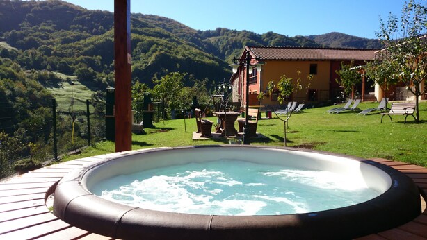 A hot tub on a wooden deck with a mountain view, a house with a red roof, and a grassy area with lounge chairs. - Apartamentos Rurales Balcón Real (Teverga)