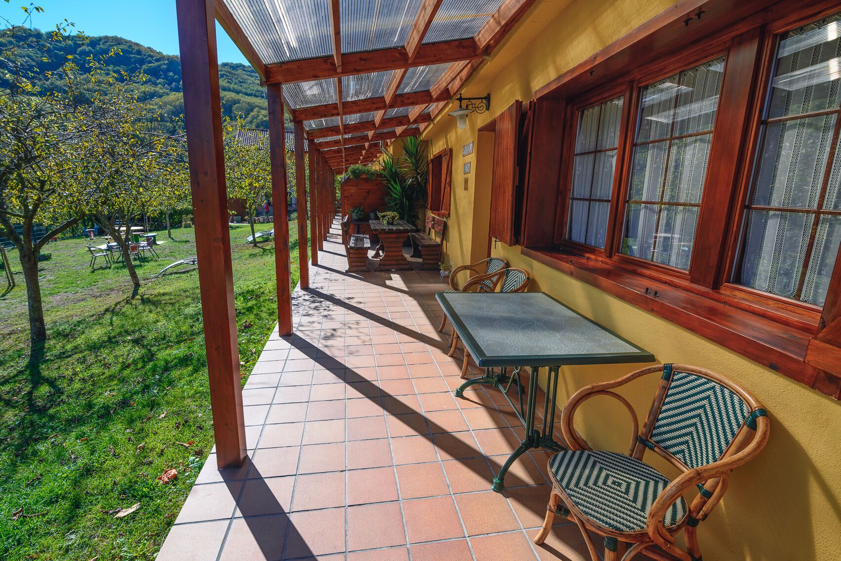 A covered patio with a table and chairs, a view of a green landscape, and a building with wooden beams and windows.