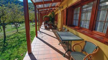 A covered patio with a table and chairs, a view of a green landscape, and a building with wooden beams and windows.