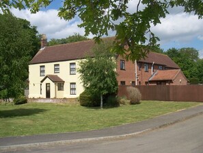 Une maison à deux étages, avec une façade en briques et un toit recouvert de tuiles, entourée d’une pelouse bien entretenue et d’une clôture en bois.