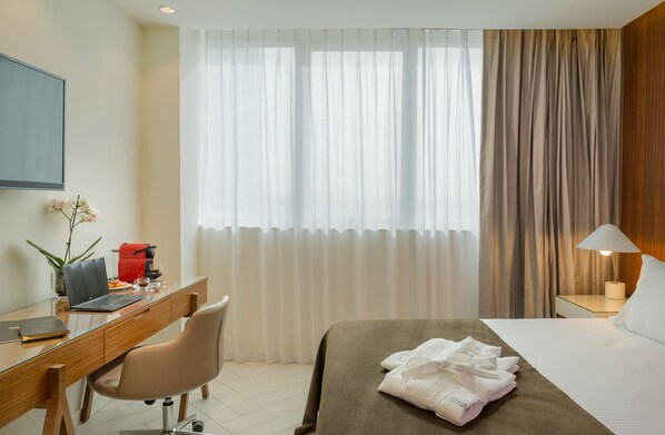 A hotel room with a bed, a desk with a laptop, a chair, a bedside table with a lamp, and a window with curtains. - Prima Millennium (Ra'ananna)